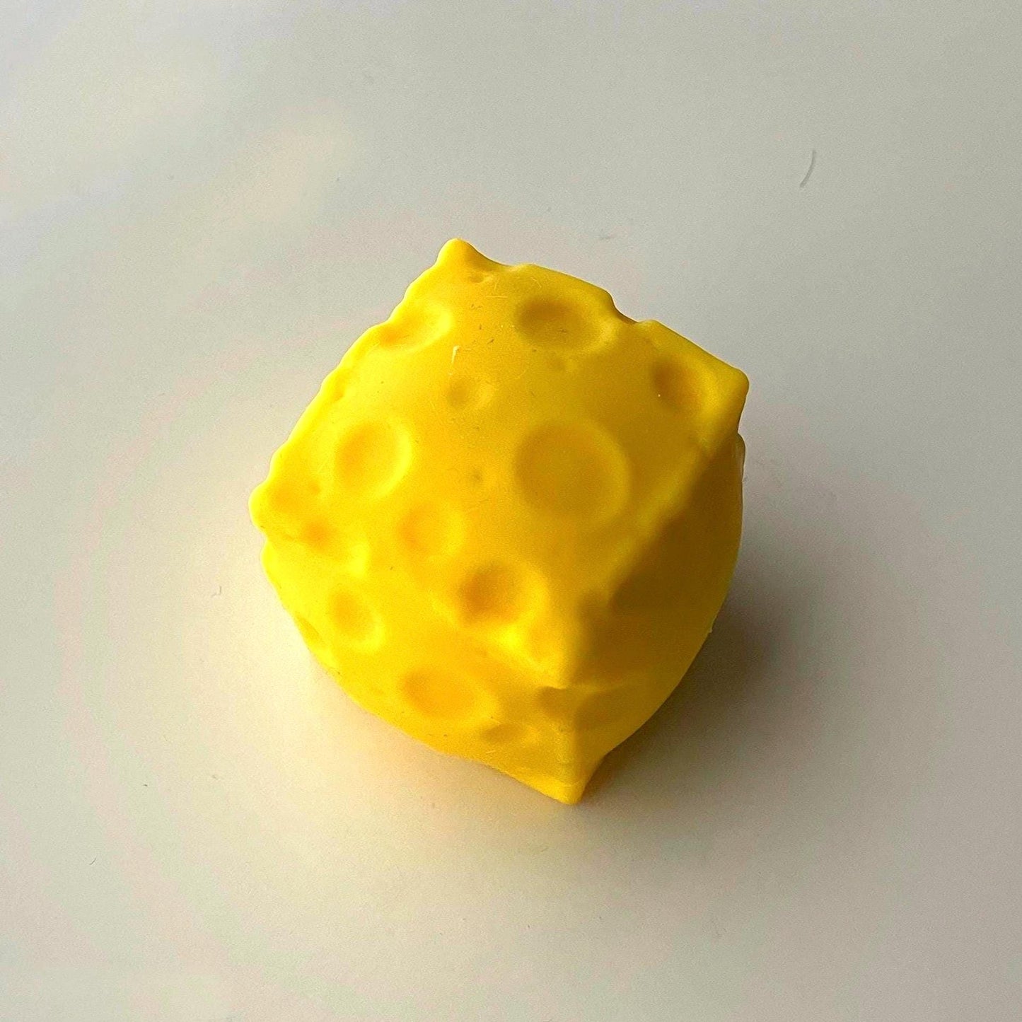 A bright yellow, cube-shaped cheese. The cheese has several round holes of varying sizes and is set against a white background. The cheese is a solid color and has a smooth texture.