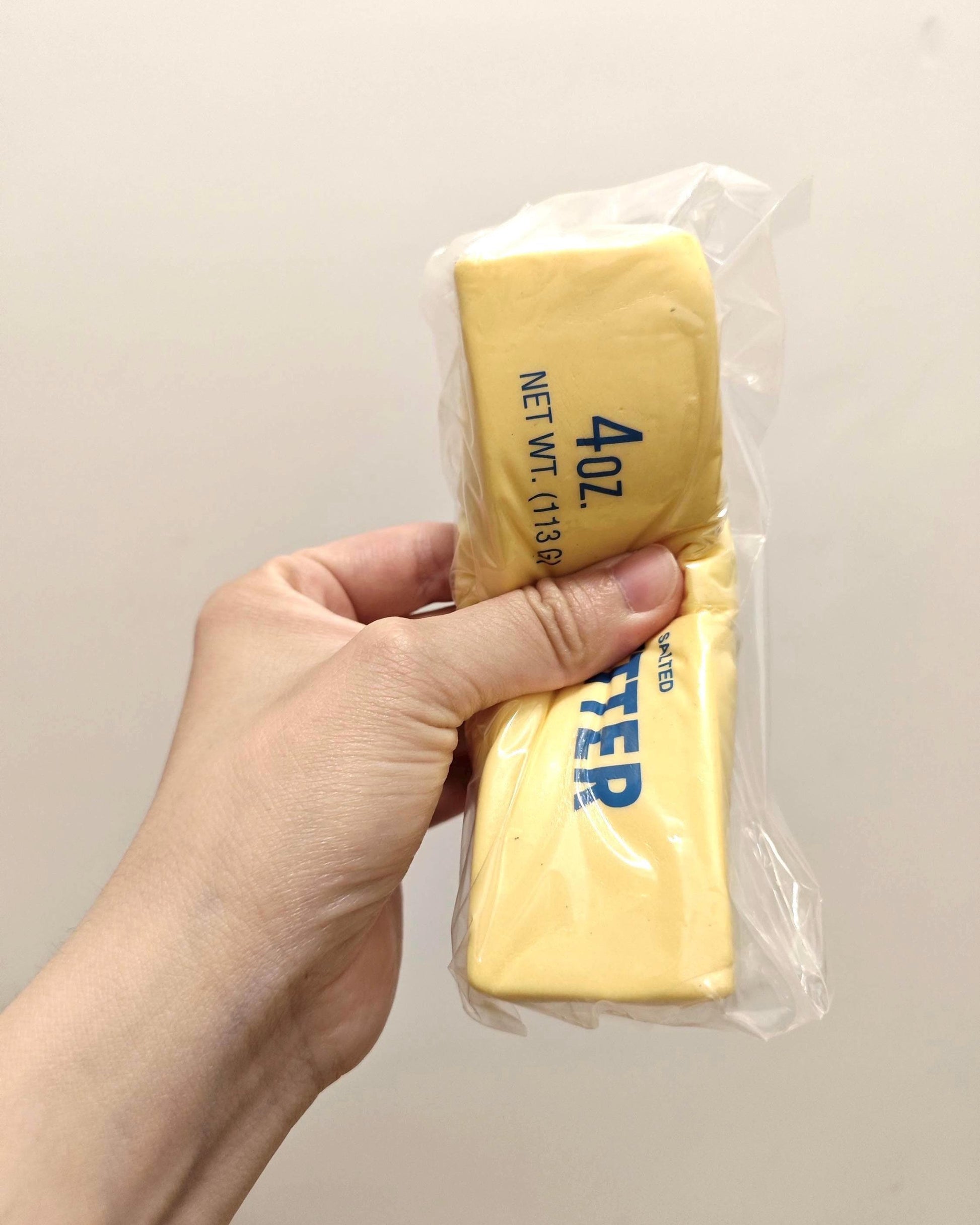 A hand holding a 4.0 lb block of butter in clear plastic wrapping against a plain off-white background.