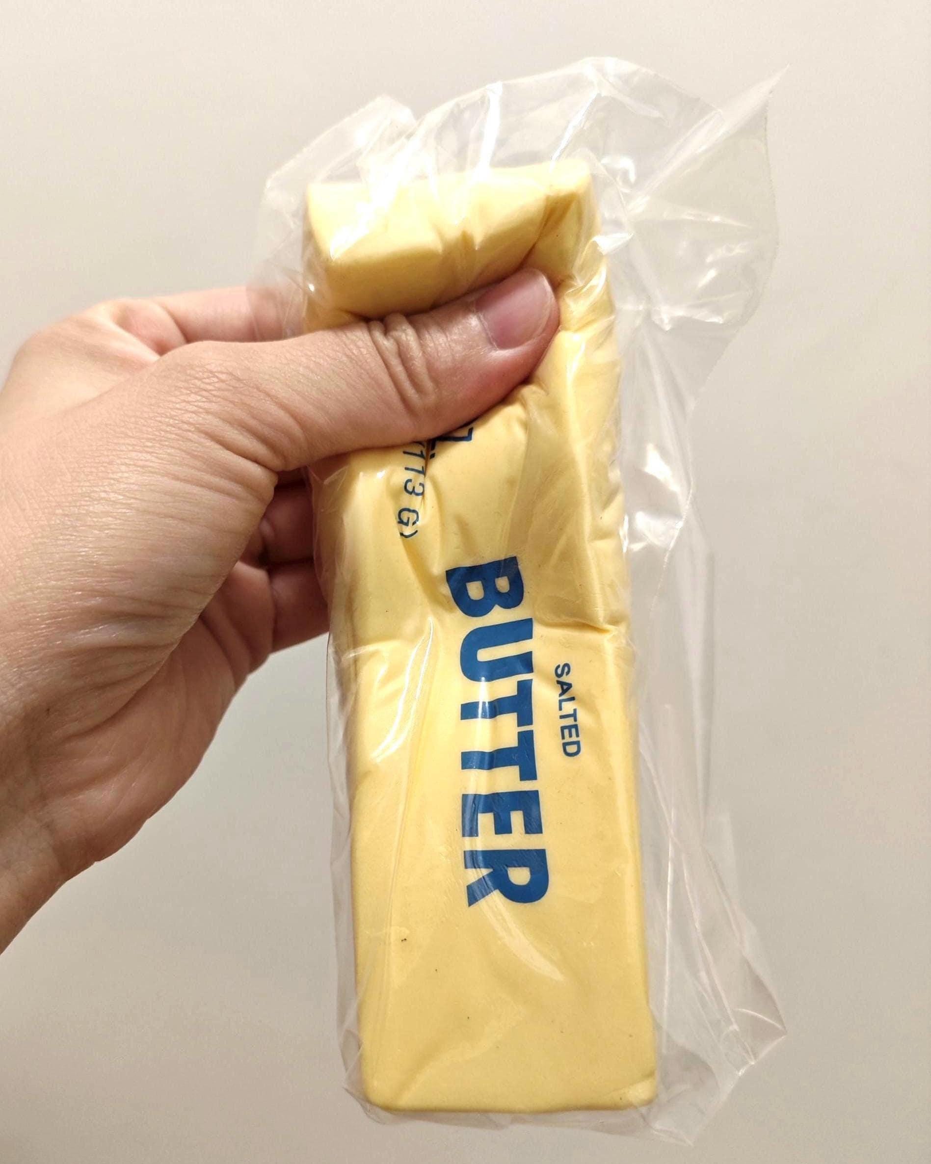 A hand holding a stick of butter in its clear plastic wrapper against a plain light background.