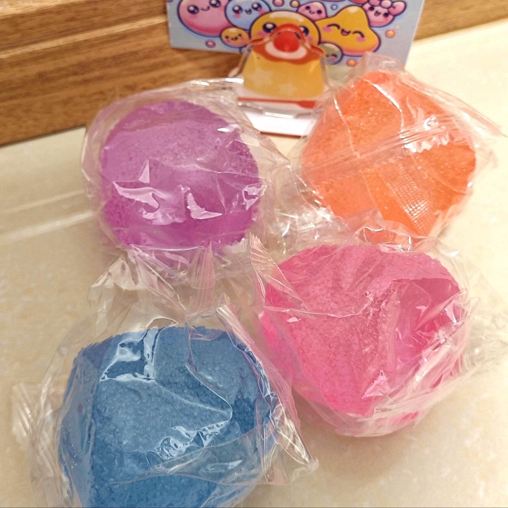 May include: Four colorful stress balls in clear plastic packaging. The balls are in shades of blue, pink, orange, and purple. A small card with the text "STRESS FREE SQUEEZE" and cartoon graphics is in the background.
