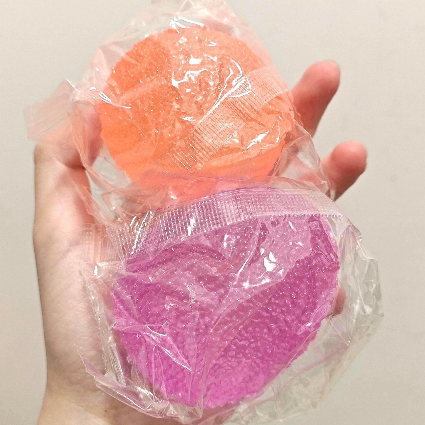 Hand holding two colorful bath bombs wrapped in plastic against a plain background