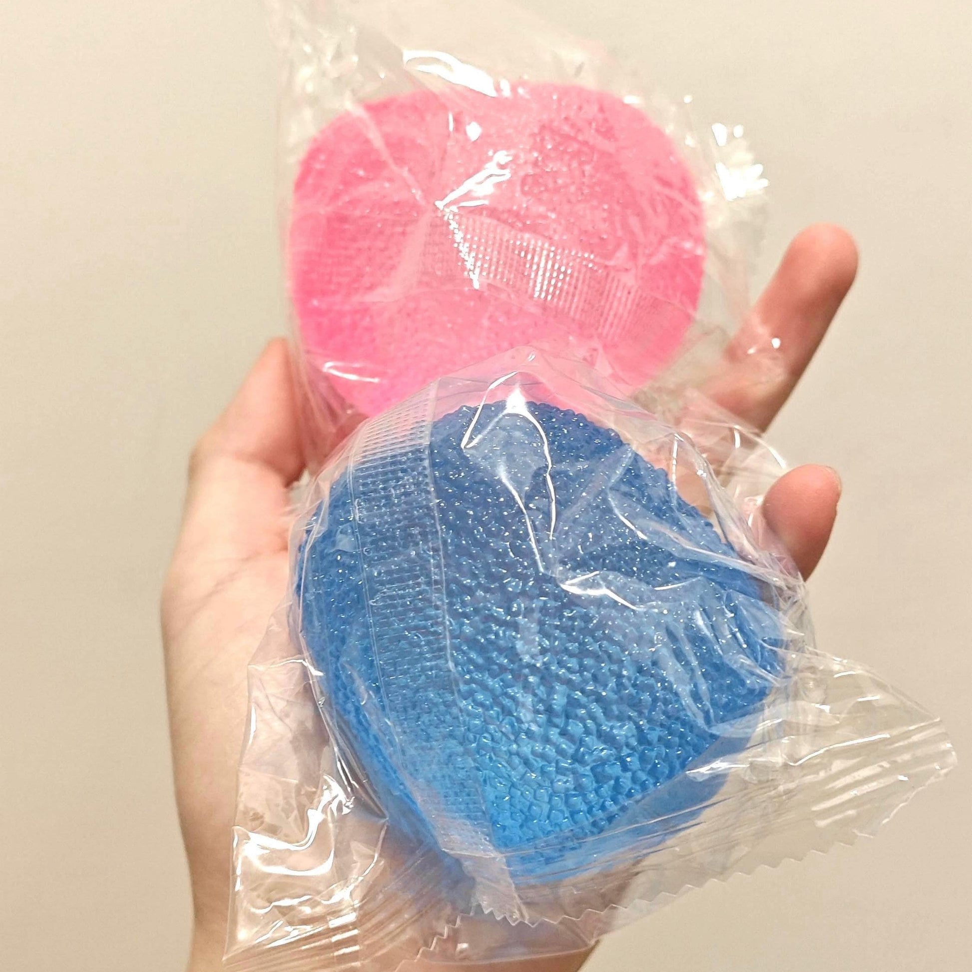 Hand holding two plastic balls, one blue and one pink, in clear packaging against a beige background.