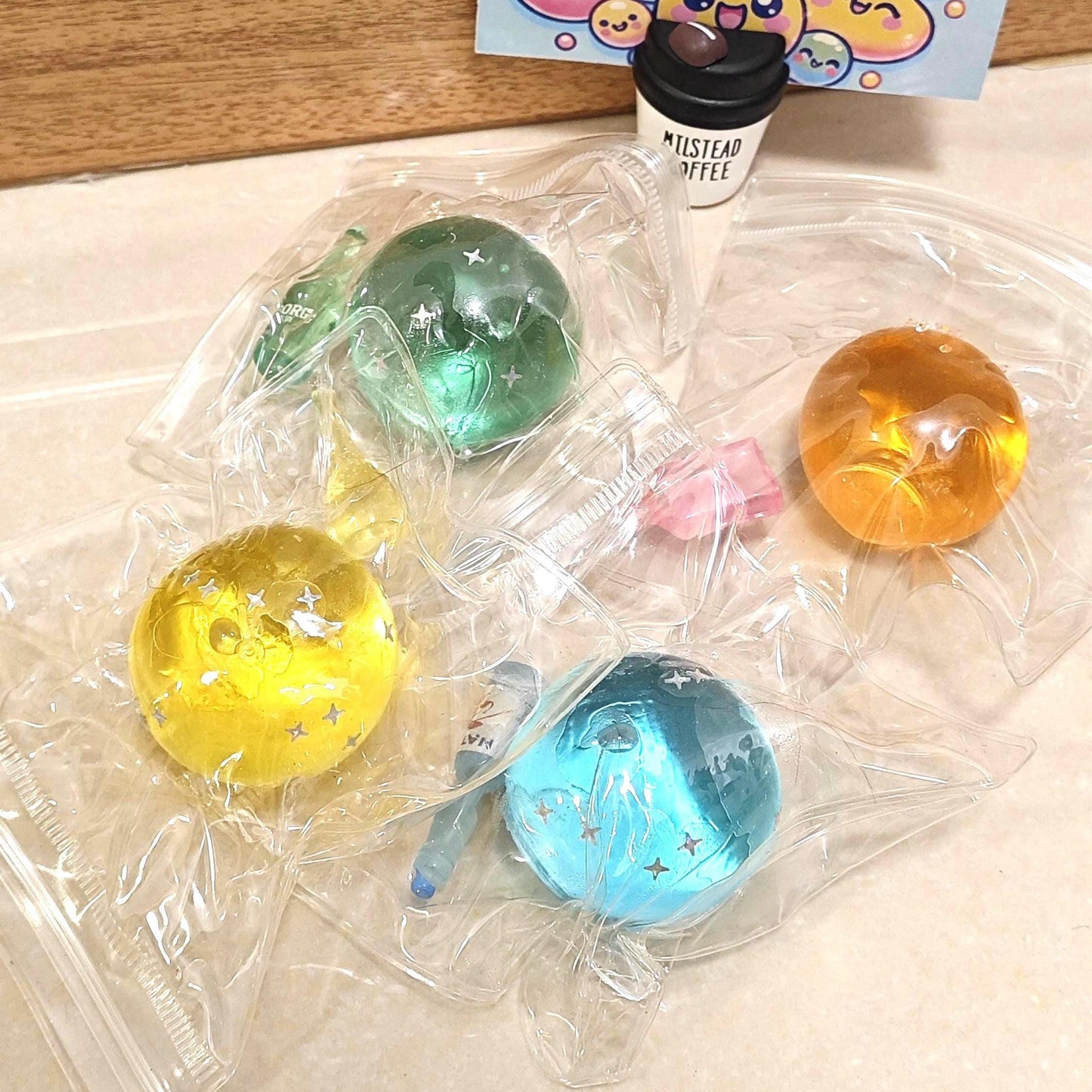 May include: An assortment of colorful, spherical stress balls in clear plastic packaging. The balls are yellow, green, orange, and blue, with star-shaped accents. A small coffee cup figurine and a card with the words "STRESS FREE SQUEEZE" are also visible.