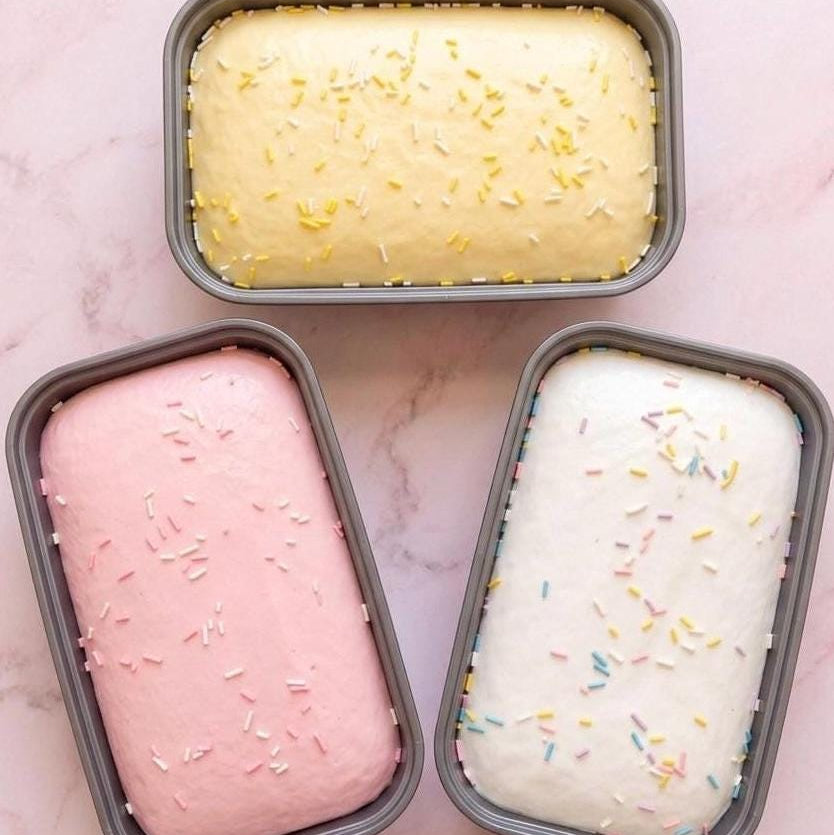 Three loaf pans with different colored cake batter and sprinkles on a pink marble background