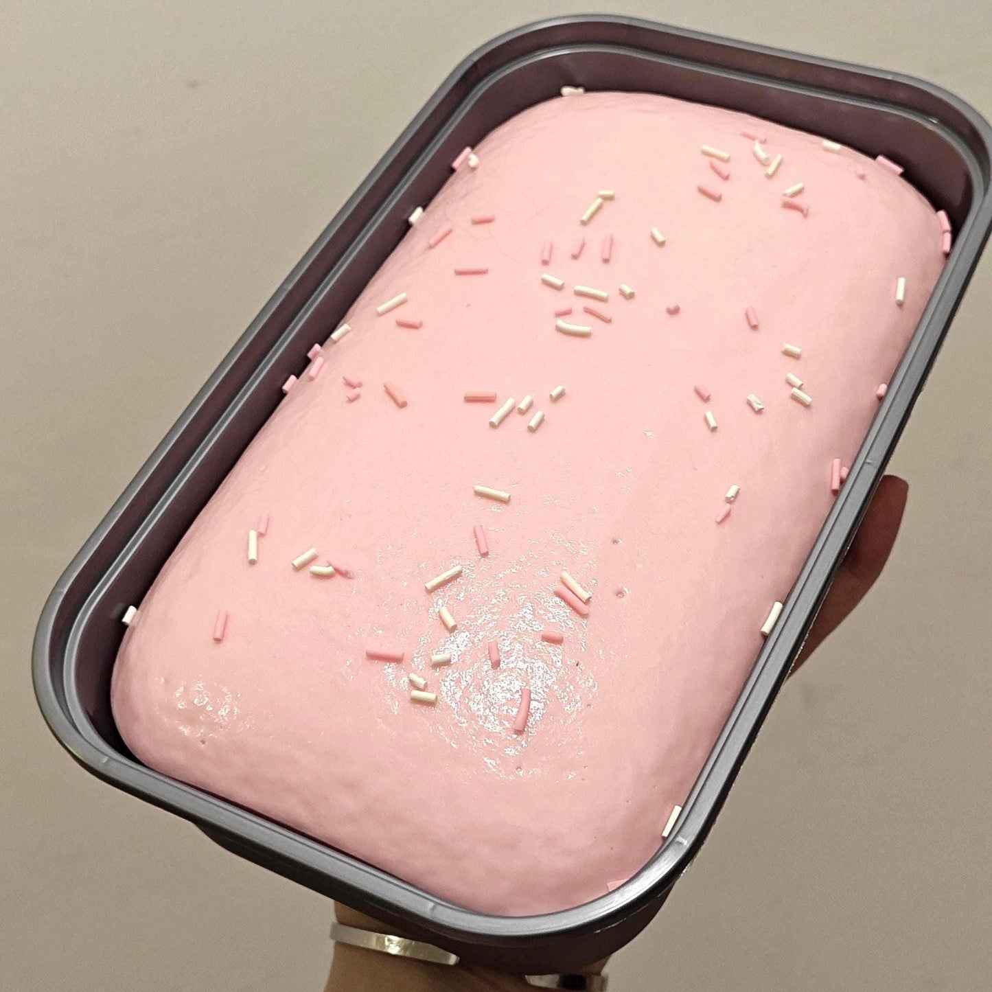 Pink ice cream loaf with sprinkles in a metal container held by a hand against a neutral background