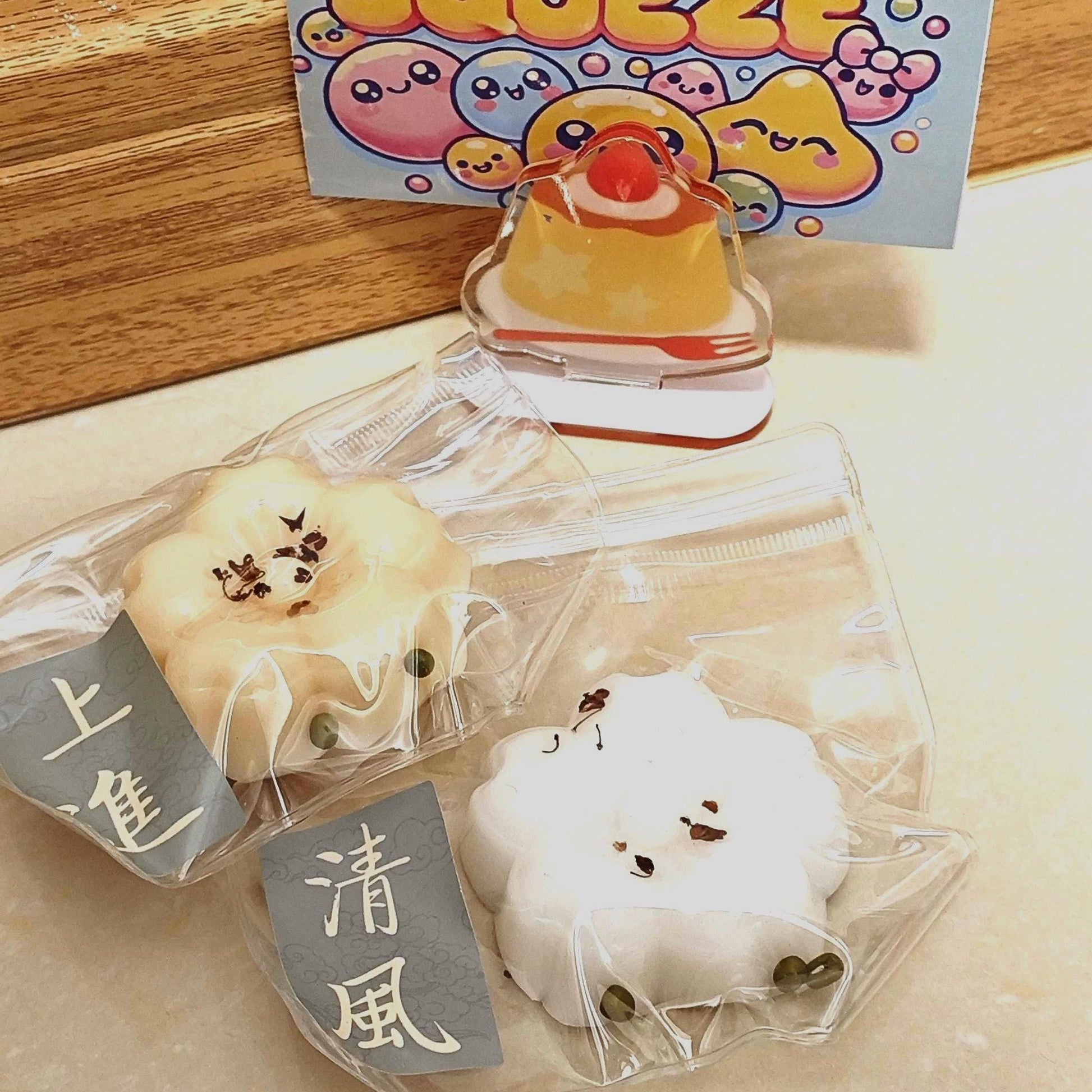 May include: A light blue card with the words "STRESS FREE SQUEEZE" in colorful bubble letters. Below the card is a clear acrylic charm shaped like a dessert. Two flower-shaped, packaged items are in the foreground.