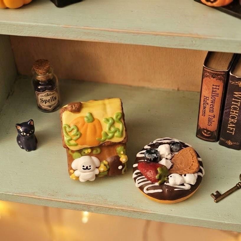 A styled shelf display featuring a small decorative gingerbread house figurine with a pumpkin roof and white ghost detail, next to a chocolate donut decorated with strawberries, blueberries, marshmallows, and a cookie, with miniature props including a black cat figurine, a bottle of spells, and books on a pale green shelf.