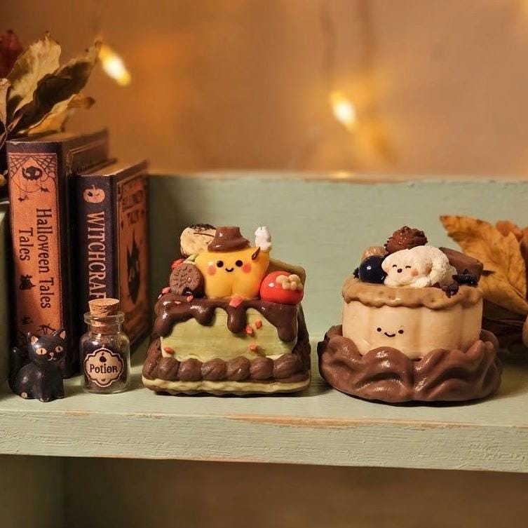 Two cute, hand-painted ceramic miniature dessert figurines displayed on a rustic light green shelf next to decorative books and a small potion bottle. The left figurine is a slice of cake with a yellow character wearing a cowboy hat, and the right figurine is a cupcake with a white character on top.