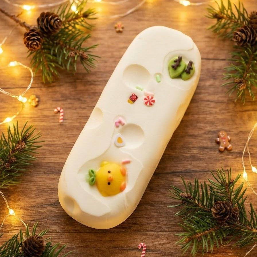 A white rectangular soap bar with cute holiday-themed illustrations, including a yellow duckling, pinecones, candy canes, and green leaves, displayed on a wooden surface decorated with pine branches, pinecones, and warm fairy lights.