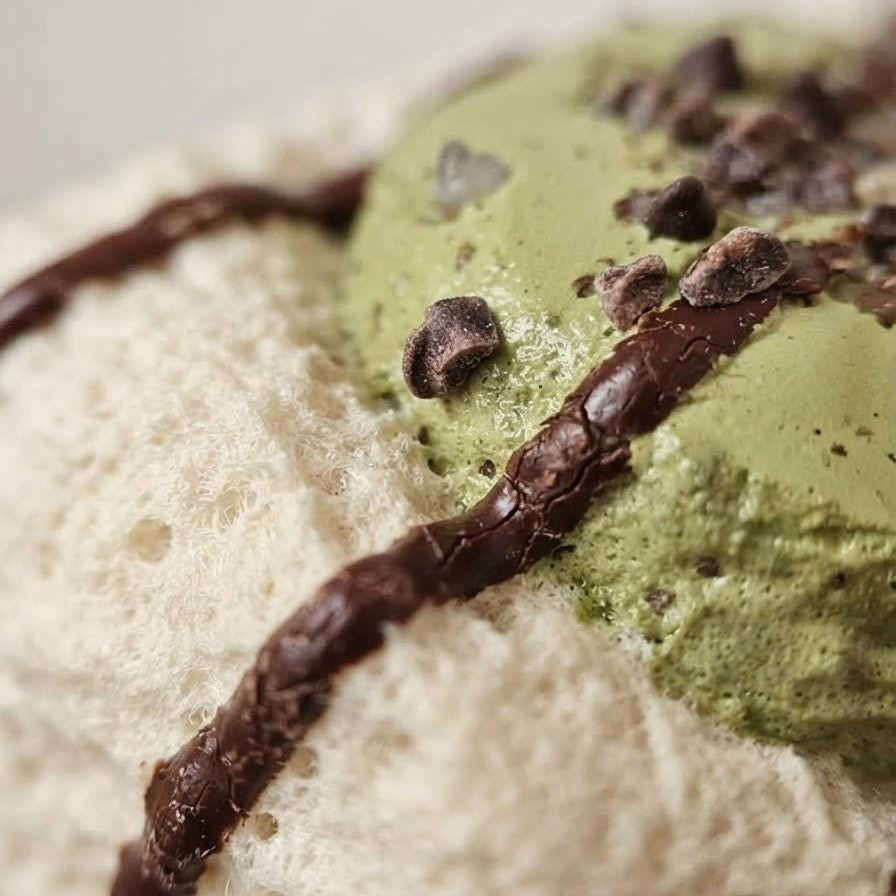 A close-up of a dessert, featuring a scoop of light green matcha ice cream topped with chocolate shavings and drizzles of chocolate sauce on a fluffy white meringue or whipped cream base.