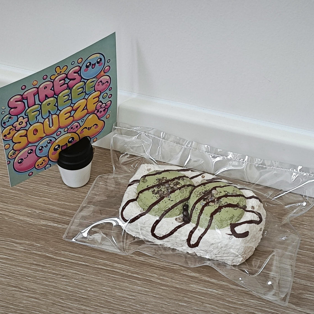 A novelty stress toy shaped like a matcha cake in a clear plastic bag, displayed on a light wood floor next to a colorful 'STRESS FREE SQUEEZE' card and a small cup.