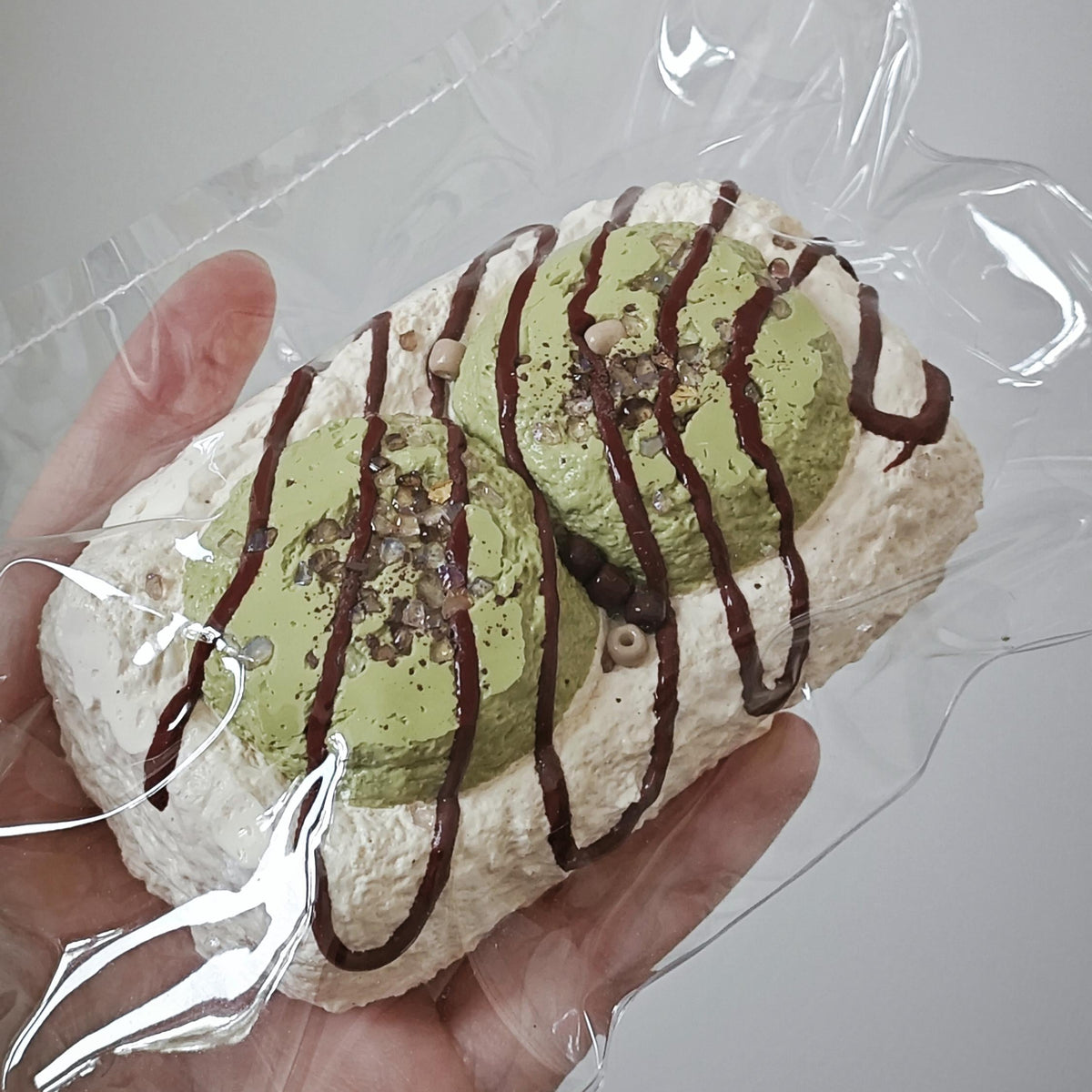 A hand holding a packaged dessert, likely a matcha-flavored pastry or cream puff, wrapped in clear plastic. The dessert has a light green matcha filling or coating, topped with dark chocolate drizzle and small chocolate chips, set against a plain light gray background.