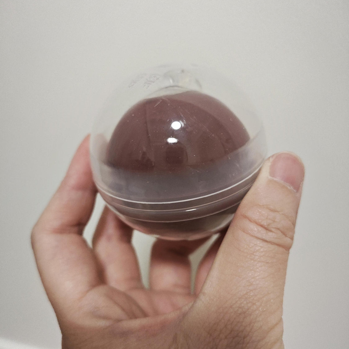 A hand holding a small, round, clear plastic capsule containing a dark brown, dome-shaped item, likely a lip balm or cosmetic product, against a plain light gray background.