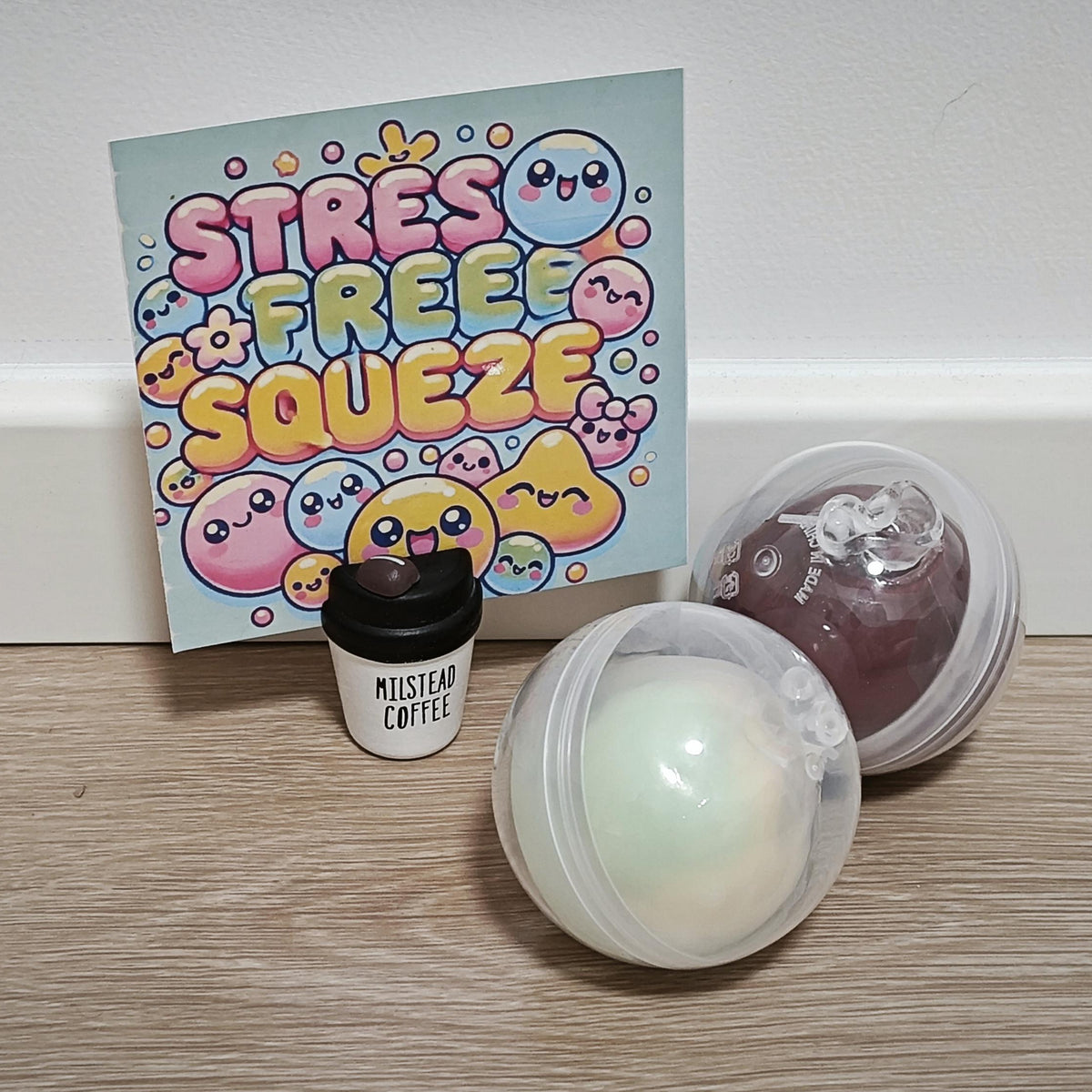 Three stress relief items arranged on a light wood floor against a white wall: a colorful card reading 'STRESS FREE SQUEEZE' with cartoon characters, a small black coffee cup labeled 'MUSTARD COFFEE,' and two translucent capsules containing round gel-like balls, one pale mint and one dark plum.