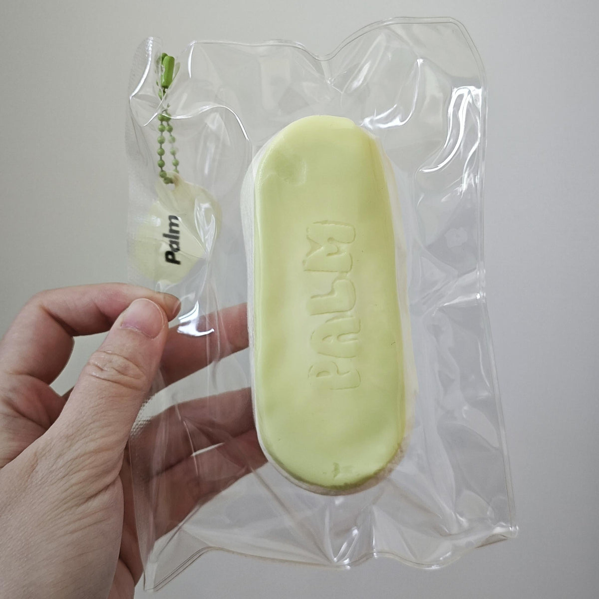 A hand holding a clear plastic bag containing a pale yellow oval-shaped soap bar with the brand name 'PALM' embossed on it, against a plain light gray background.