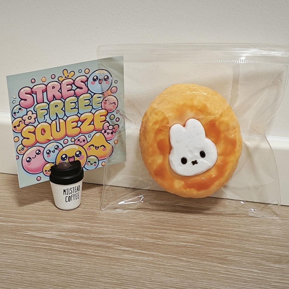 A round orange and yellow textured squishy toy with a white bunny face, sealed in a clear plastic bag next to a small coffee cup accessory and a colorful 'Stress Free Squeeze' card, set on a light wood surface against a white wall.
