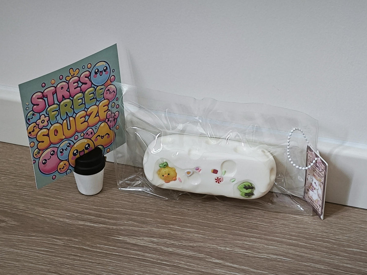 A white silicone stress relief toy with cute cartoon graphics, shown in its clear plastic packaging next to a small black travel cup and a colorful 'STRESS FREE SQUEEZE' card on a wooden floor against a white wall.