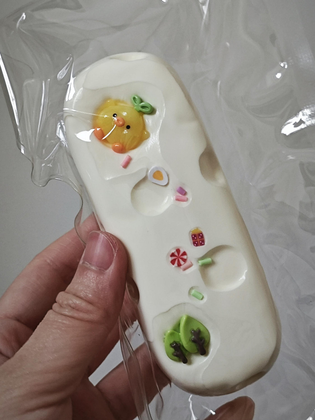 A hand holding a white, elongated novelty item wrapped in clear plastic. The item has molded holes and small colorful decorative details, including a yellow duck face near the top and green leaf shapes near the bottom.