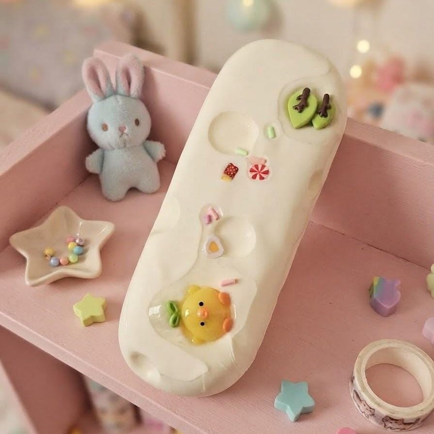 A pastel pink and white decorative phone case with cute cartoon embellishments, displayed on a pink shelf with a small plush bunny, star-shaped dish, and other pastel accessories.