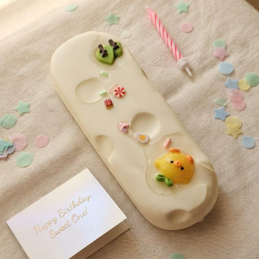 A cream-colored silicone bath mat with cute animal and flower details, styled on a beige fabric surface with a pink candle, star confetti, and a small birthday card that reads 'Happy Birthday Sweet Oral!'.