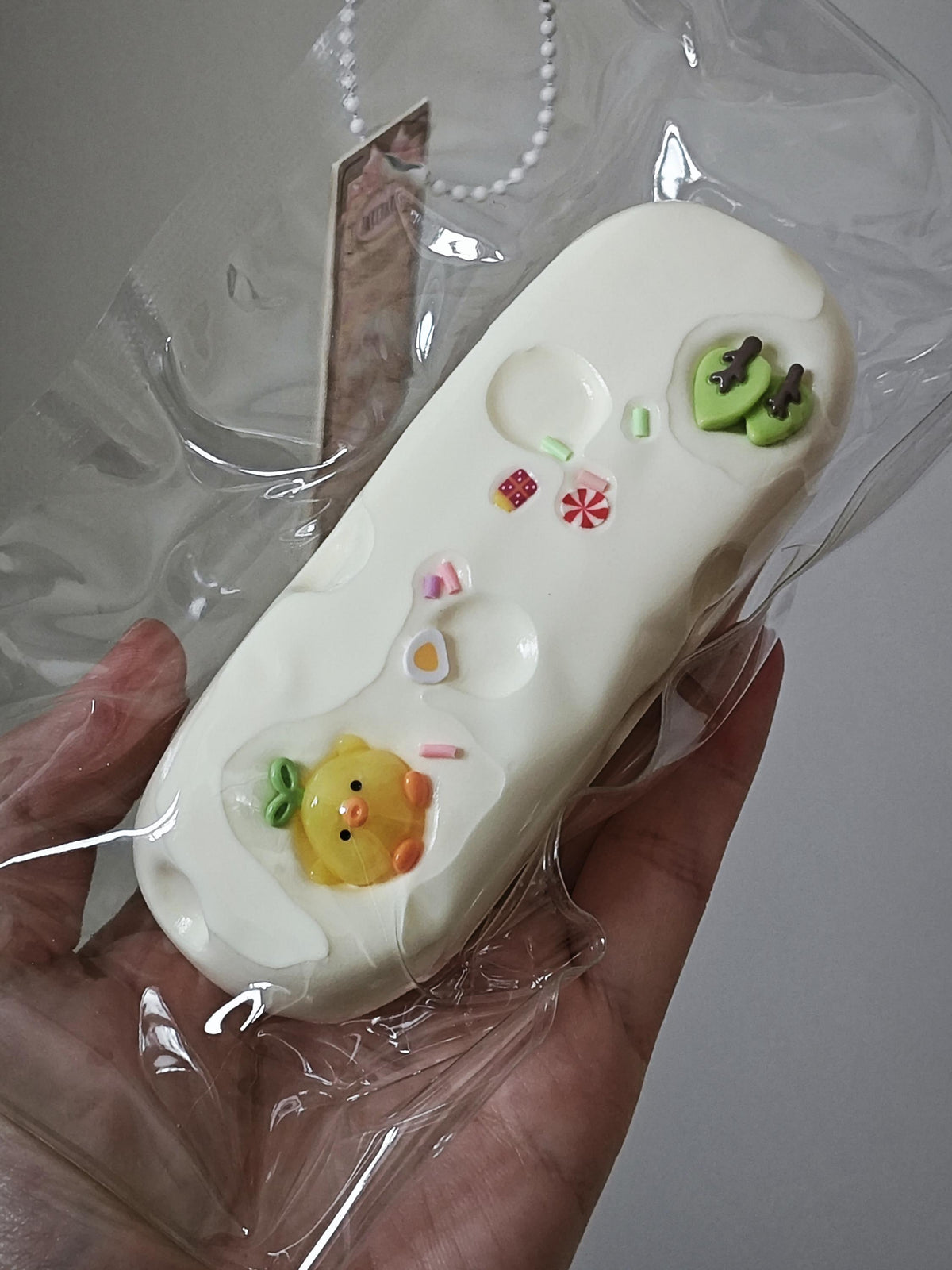 A hand holding a white silicone bath bomb mold in clear plastic wrapping, decorated with a yellow chick, green kiwi, and small candy details.