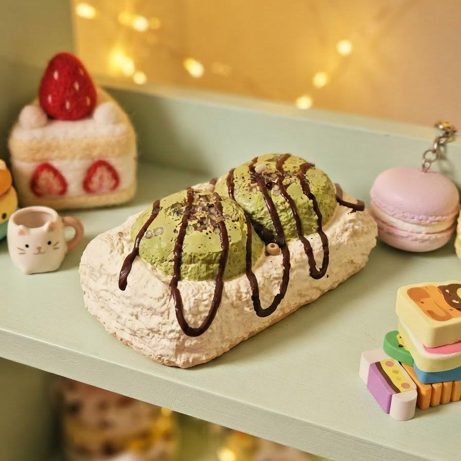 A close-up of a miniature toy dessert pastry with green matcha-like filling and dark chocolate drizzle, displayed on a light green shelf with other cute toy food items and warm string lights in the background.