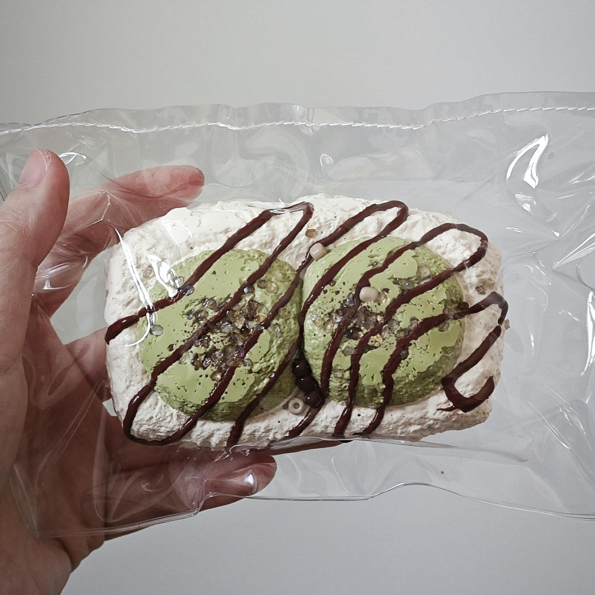 A hand holding a clear plastic bag containing two round green pastries topped with dark chocolate drizzle, against a plain light gray background.