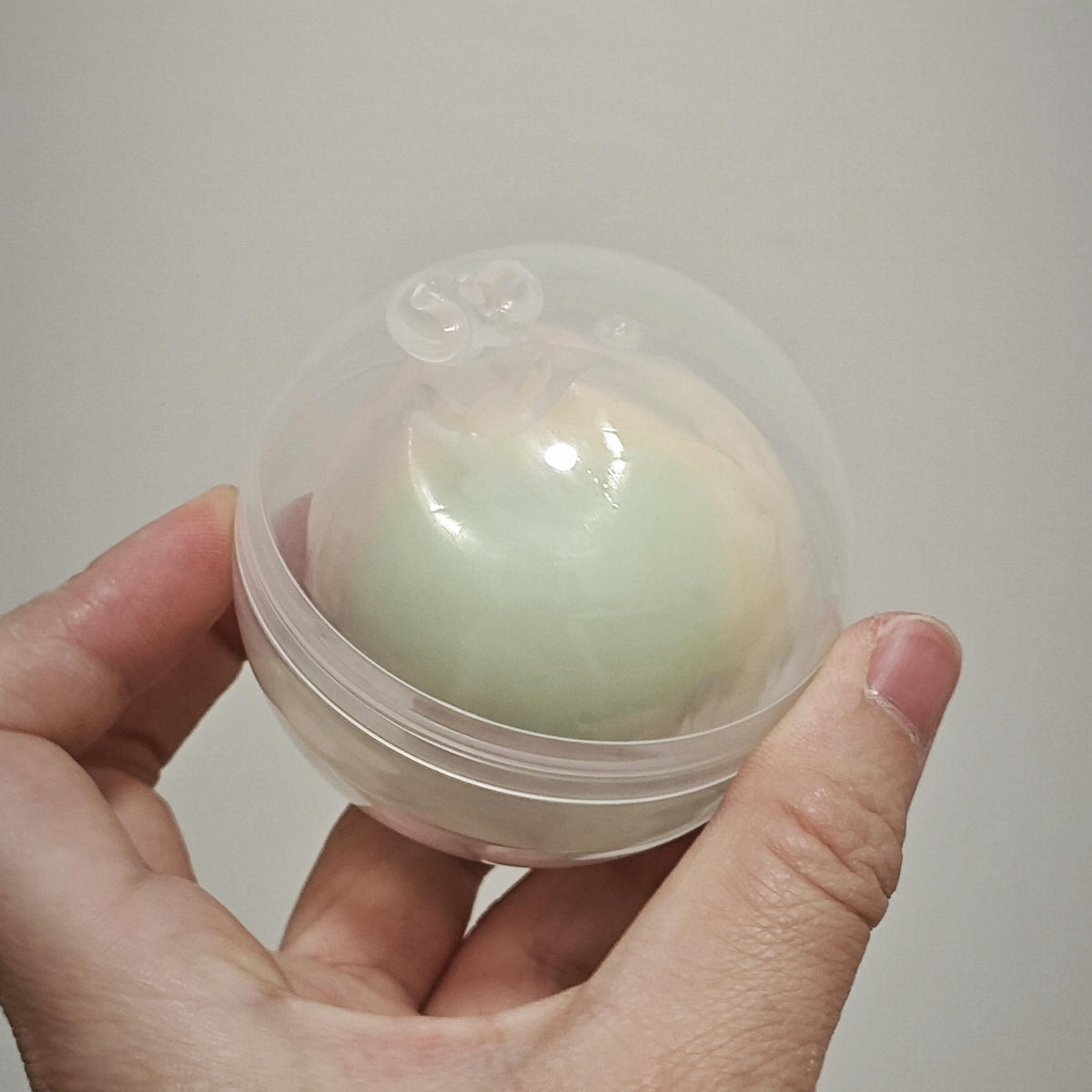 A hand holding a small clear plastic capsule containing a pale mint-green oval item against a plain light gray background.