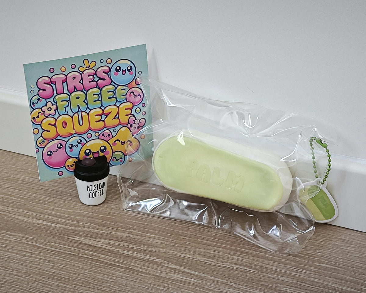 A pale yellow-green squishy stress toy in clear plastic packaging, displayed next to a small cup-shaped product labeled 'MUST SEE COFFEE' and a colorful card that reads 'STRESS FREE SQUEEZE'.