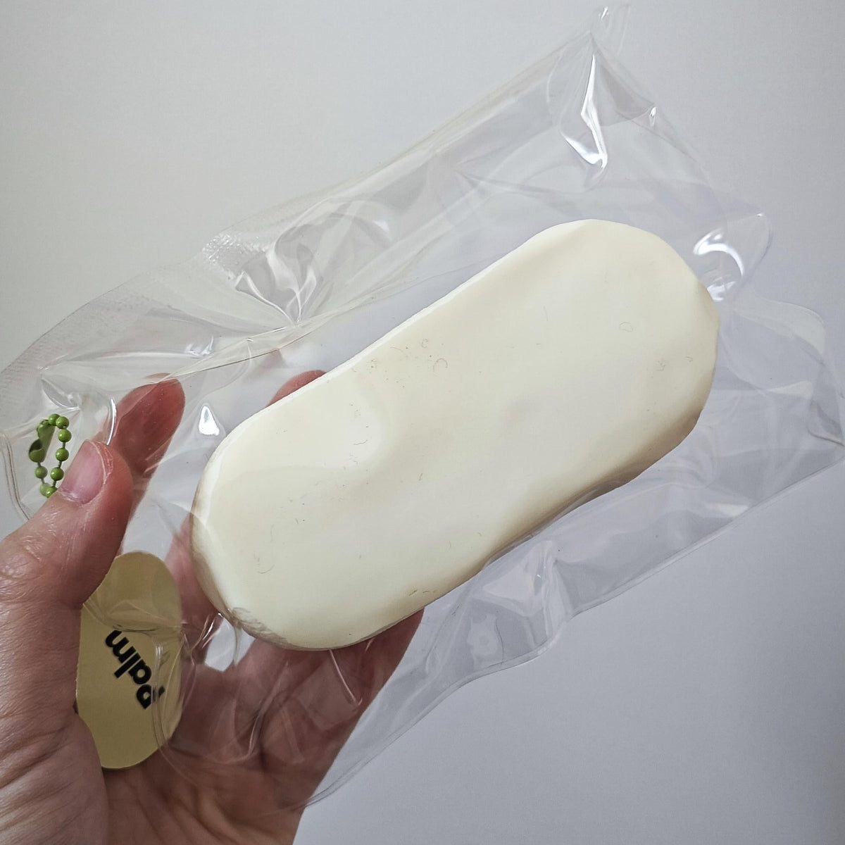 A hand holding a clear plastic bag containing a smooth, off-white oval-shaped item, with a small gold tag attached. The background is a plain light gray wall.