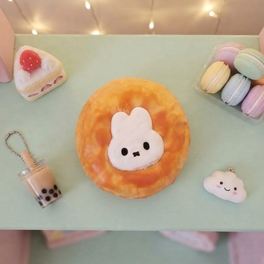 A cute, round, orange-brown donut-shaped squishy toy with a white bunny face on the front, displayed on a mint green tabletop alongside other miniature pastel accessories.