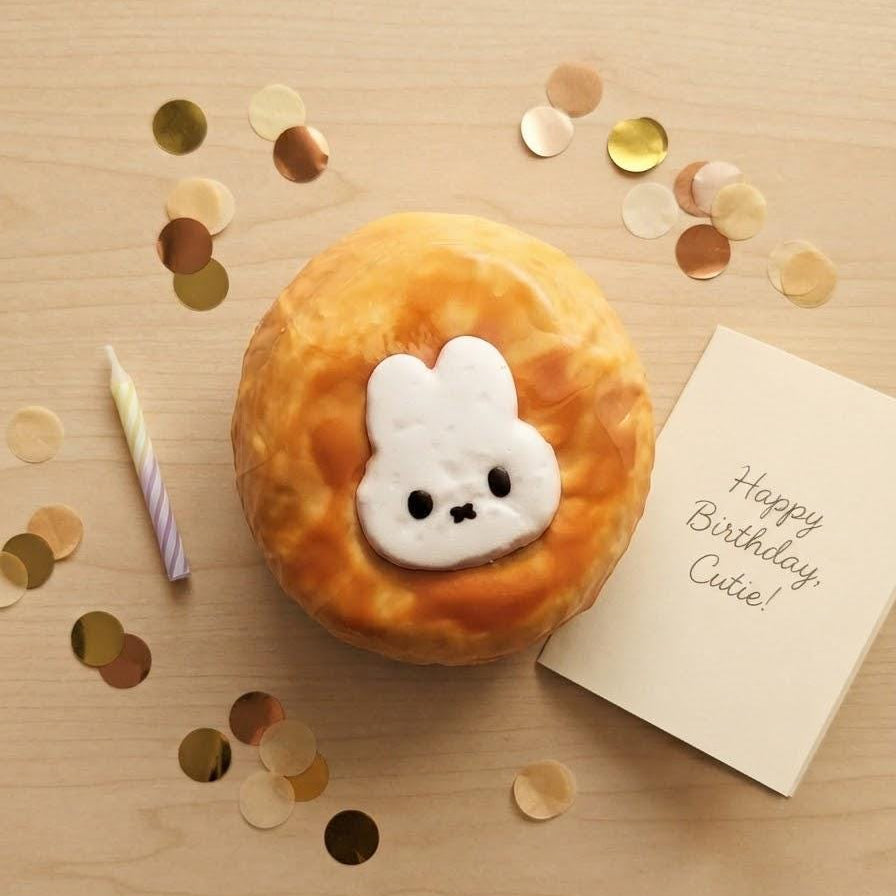 A round, golden-brown donut-shaped squishy toy with a white bunny face sits on a light wood surface, surrounded by gold confetti, a small striped candle, and a card that reads 'Happy Birthday Cute!'.