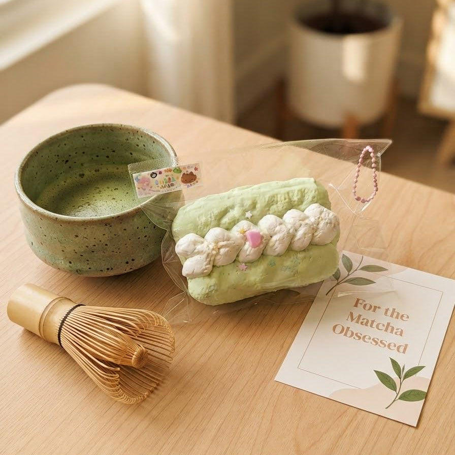 A styled flat-lay of a matcha-themed product set on a light wood table, featuring a light green rectangular matcha cake with white cream and pink details, a green speckled ceramic bowl, a bamboo whisk, and a card that reads 'For the Matcha Obsessed'.