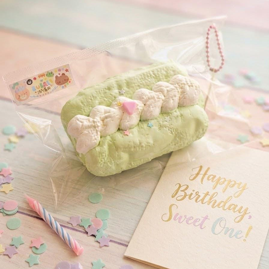 A pastel green novelty squishy toy shaped like a donut with white marshmallows and a pink bow, shown in clear plastic packaging on a light wooden surface with confetti, a candle, and a birthday card reading 'Happy Birthday, Sweet One!'.