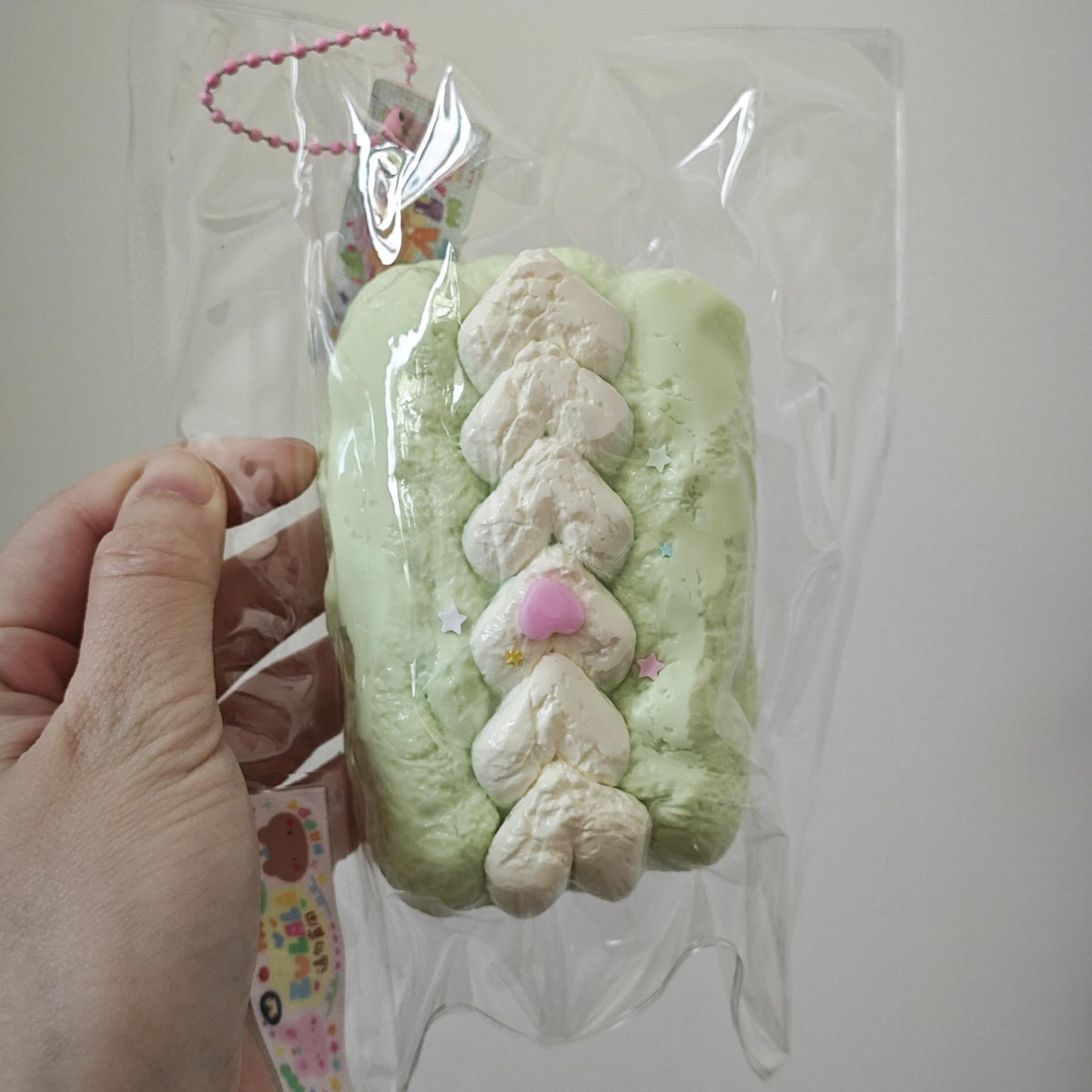 A hand holding a clear plastic bag containing a light green rectangular item with a cream-colored textured vertical design and a small pink accent, against a plain light gray background.