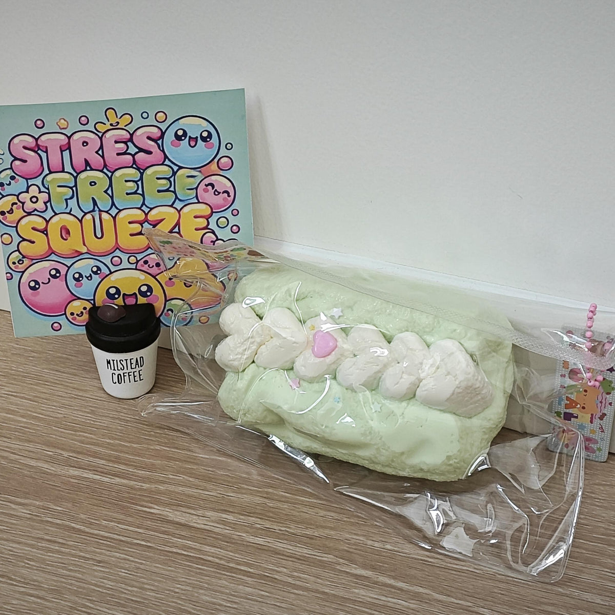 A plush squishy toy shaped like a mint green pastry with white marshmallows and a pink center, shown in clear plastic packaging on a wooden floor next to a small coffee cup and a colorful 'STRESS FREE SQUEEZE' card.