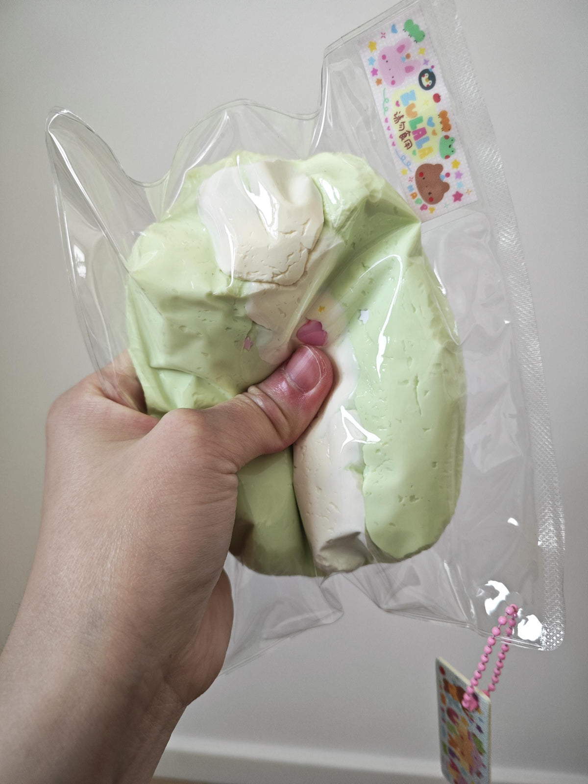 A hand holding a packaged squishy toy made from pale green and white material inside a clear plastic bag with a small attached tag.