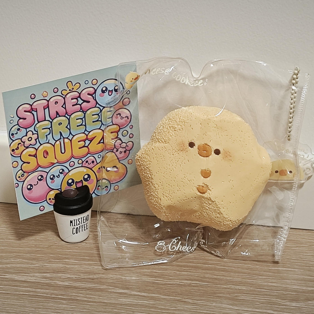 A beige, cloud-shaped squishy toy with a cute face, shown inside a clear plastic bag next to a small coffee cup accessory and a colorful card that reads 'STRESS FREE SQUEEZE'.