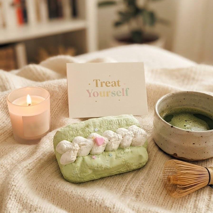 A styled cozy scene on a cream knit blanket with a lit candle, a small card reading 'Treat Yourself,' a light green pastry-shaped item with white piped frosting details, a ceramic cup of green tea, and a bamboo whisk in the background.