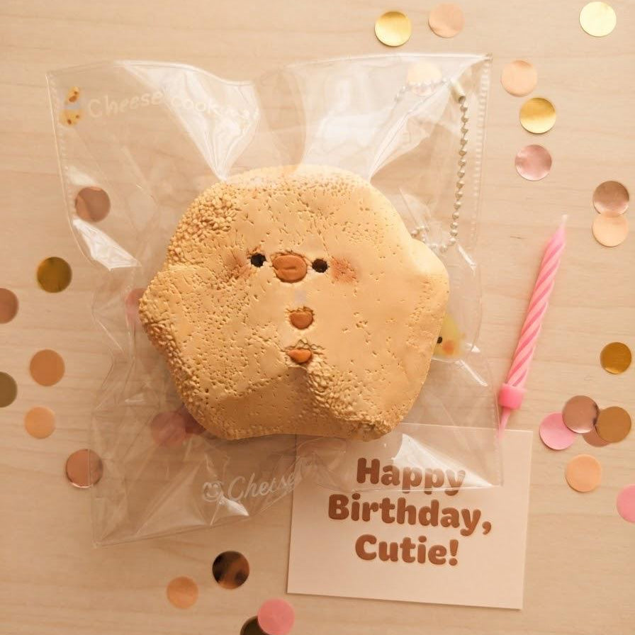 A cute, round, tan clay-like character squishy toy with a small face, sealed in a clear plastic bag. A small pink candle and a card reading 'Happy Birthday, Cutie!' are placed beside it on a light wood surface scattered with gold and pink confetti.
