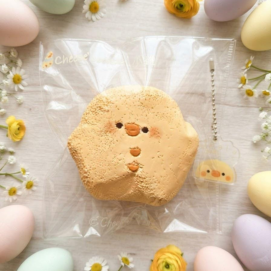 A cute, chick-shaped squishy toy in a clear plastic bag, surrounded by pastel Easter eggs, white daisies, and yellow flowers on a light wooden background.