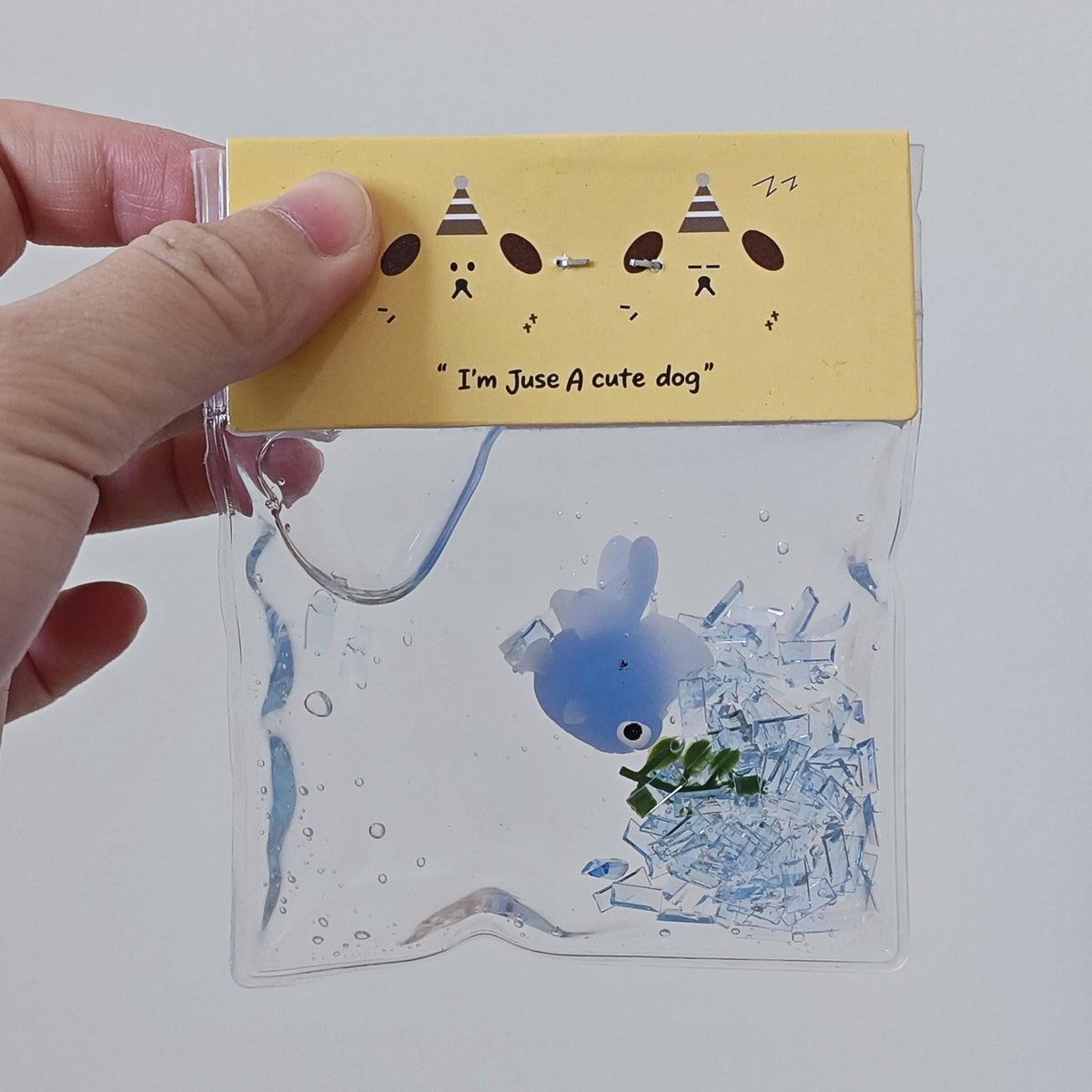 A hand holding a clear plastic bag with a yellow paper header that reads "I'm Just A cute dog" and shows a blue whale and green leaves inside.
