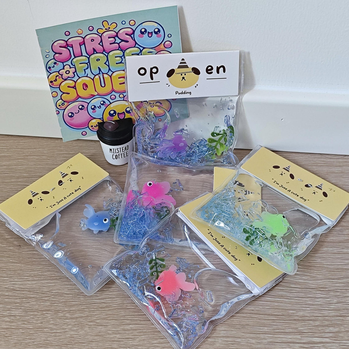 A set of colorful stress relief squishies in clear plastic bags with yellow cards, a small black jar, and a 'STRESS FREE SQUEEZE' book on a light wood floor against a white wall.