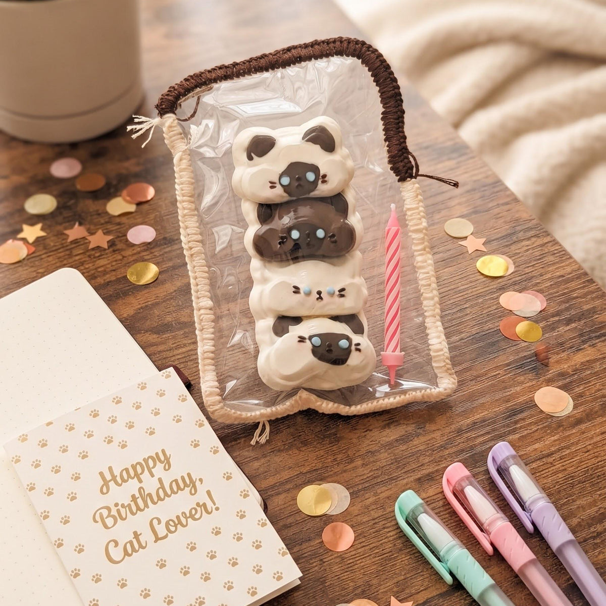 A clear zip pouch with a brown handle, filled with three stacked cat-shaped cookies and a pink candle, styled on a wooden table with a notebook, pens, and confetti.