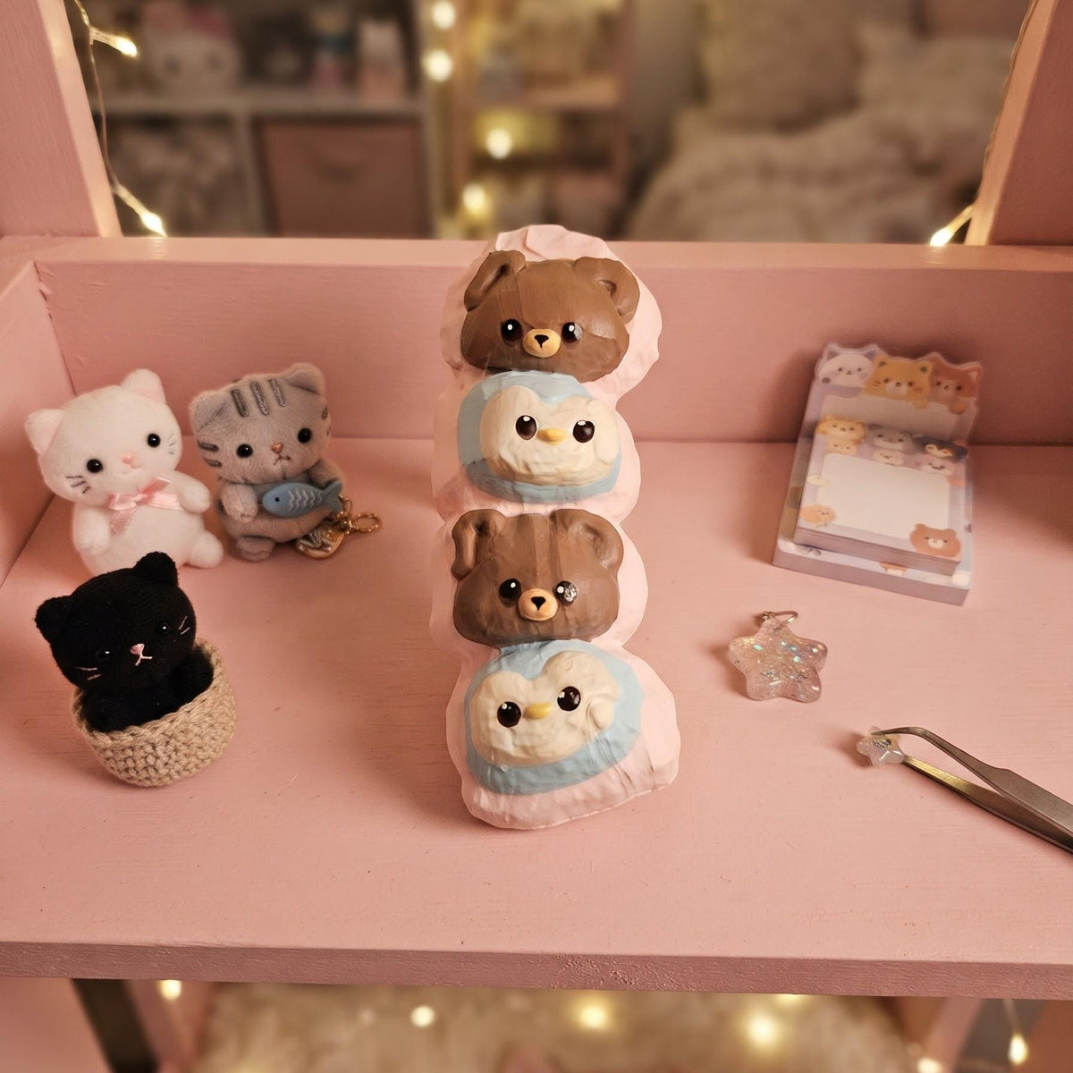 A pink miniature shelf display featuring a stack of three bear snowman figurines, small plush cat figures, a black cat in a basket, a notepad, and a star-shaped accessory, with warm fairy lights in the blurred background.