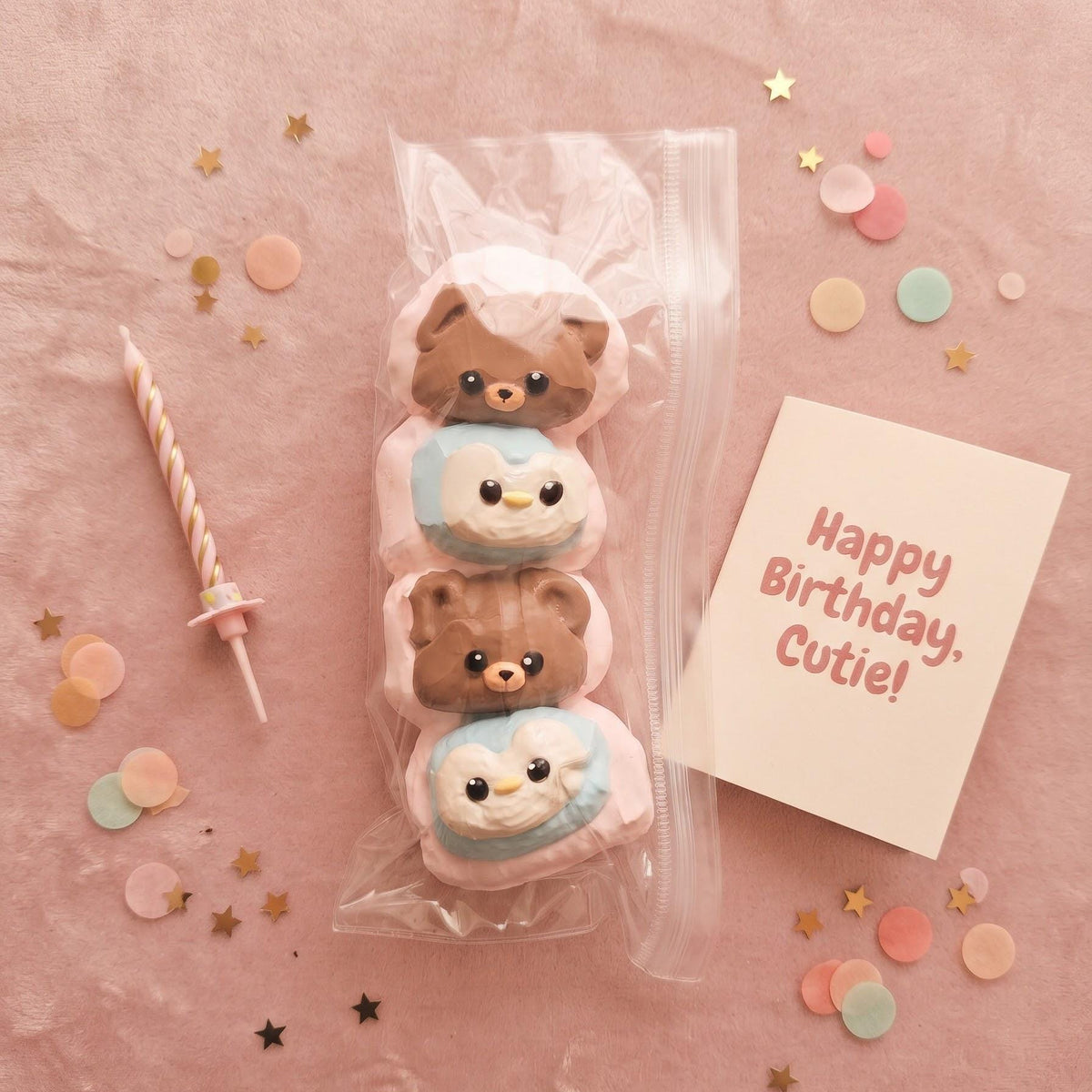 A set of four cute bear-themed birthday treats in a clear plastic bag, displayed on a pink textured background with a candle, confetti, and a 'Happy Birthday, Cutie!' card.