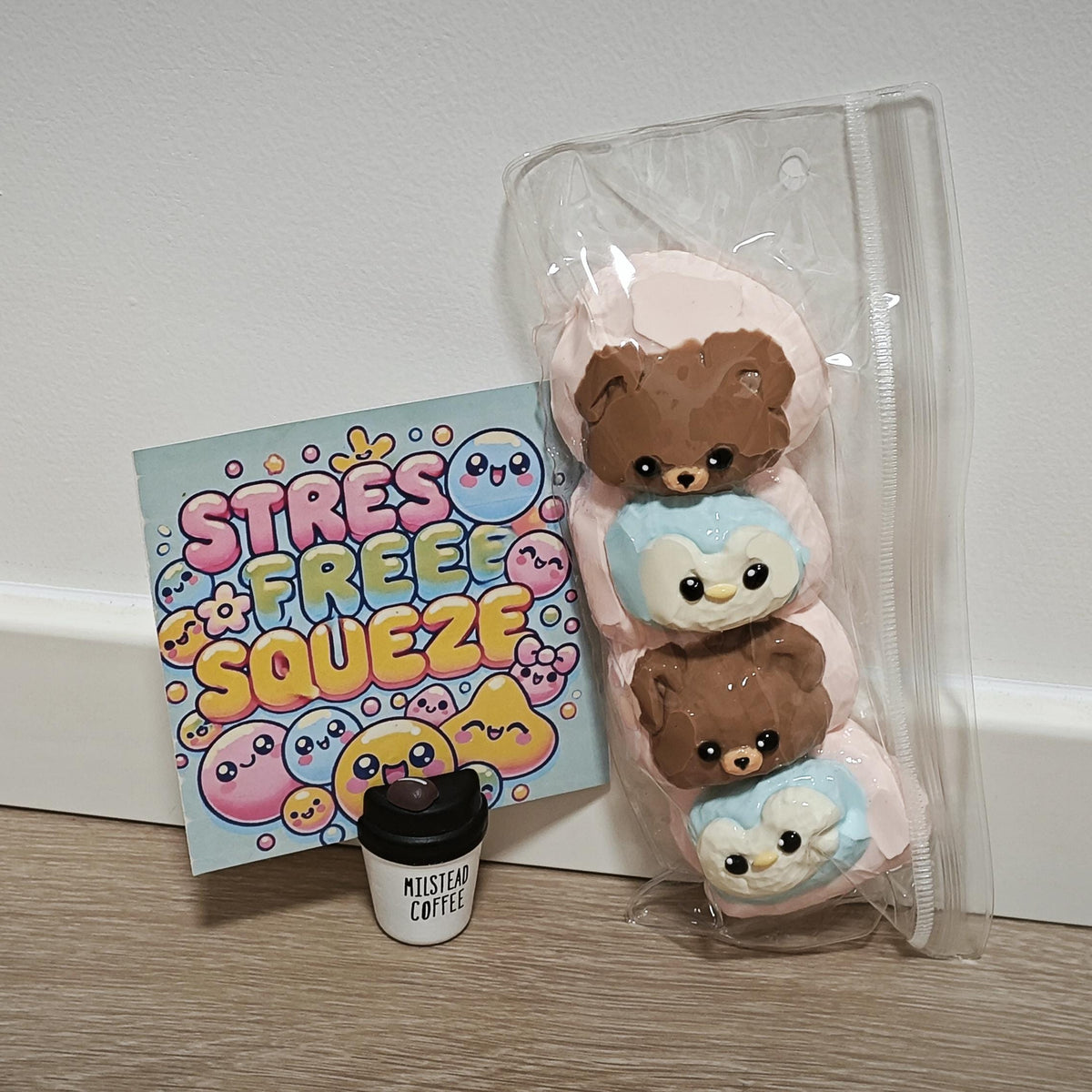 A clear plastic bag containing three small squishy plush toys shaped like teddy bears on pastel pink bunnies, shown next to a printed card that reads 'STRESS FREE SQUEEZ' and a small coffee cup accessory on a wood floor against a white wall.