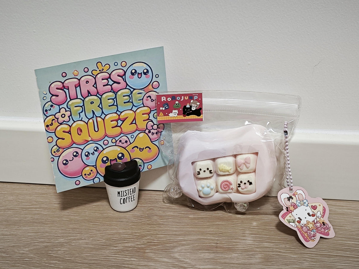 A collection of cute pastel-themed stationery and accessories arranged on a wooden surface against a white wall. The set includes a 'STRESS FREE SQUEEZE' card, a small 'Musical Coffee' cup, a pink squishy toy in a clear bag, and a Hello Kitty star keychain.