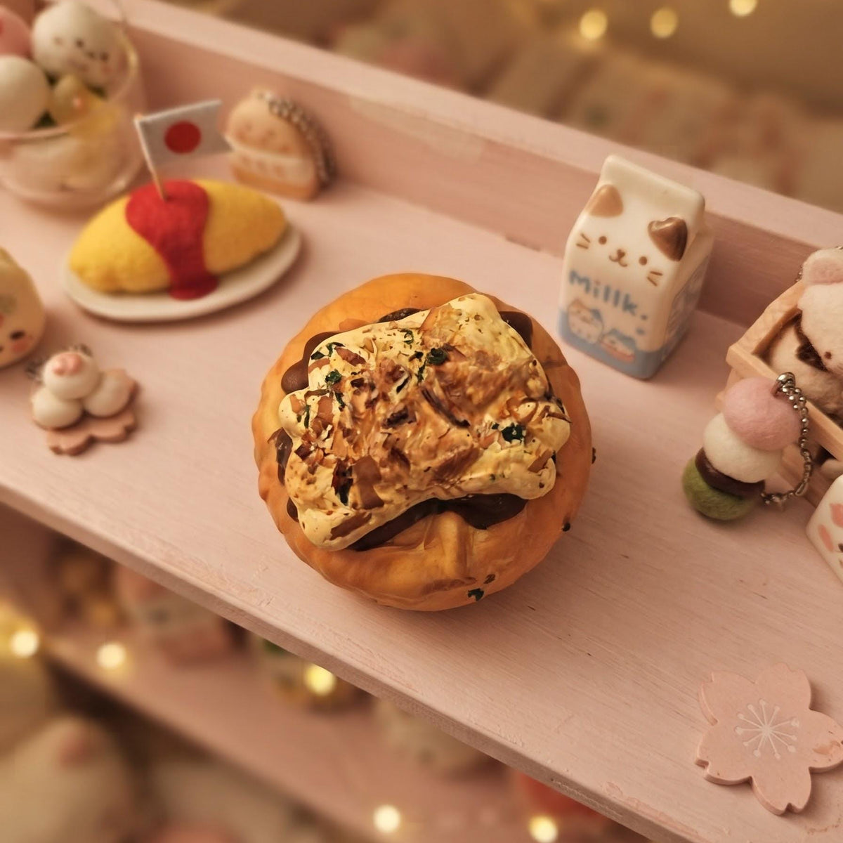A miniature toy or model of a Japanese omurice pancake sandwich with a small Japanese flag, displayed on a pink wooden shelf surrounded by cute decorative items and warm fairy lights.