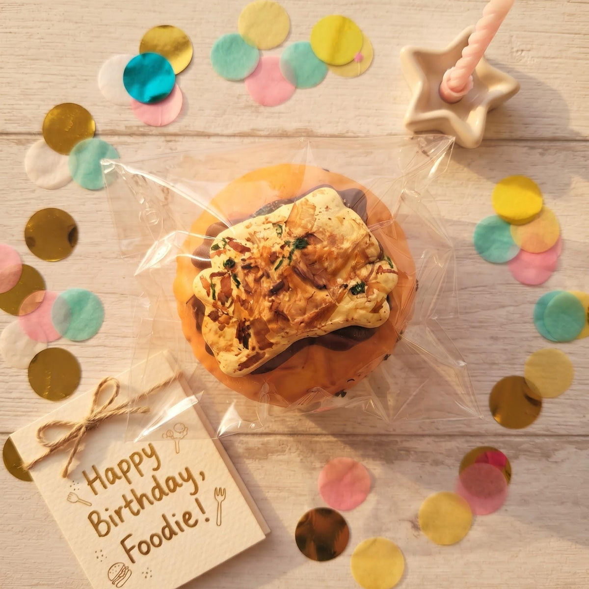 A baked good wrapped in clear cellophane sits on a light wood surface with colorful confetti, a small card that says 'Happy Birthday, Foodie!', a star candle holder, and other party-style props.